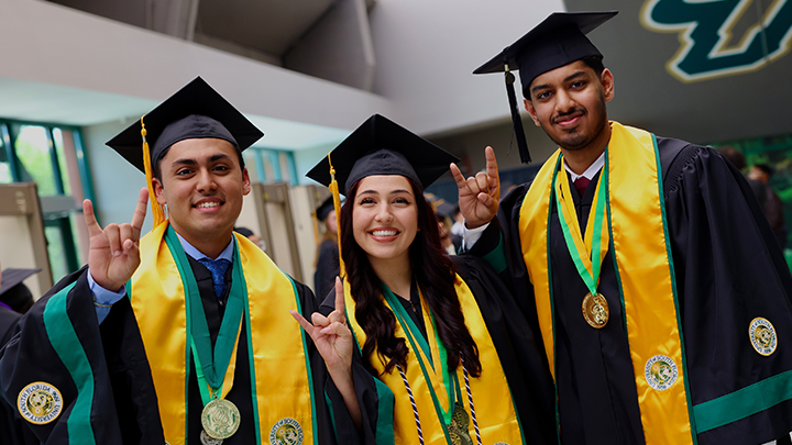 Three USF graduates wearing Commencement regalia smiling at the camera with Bull U hand signs