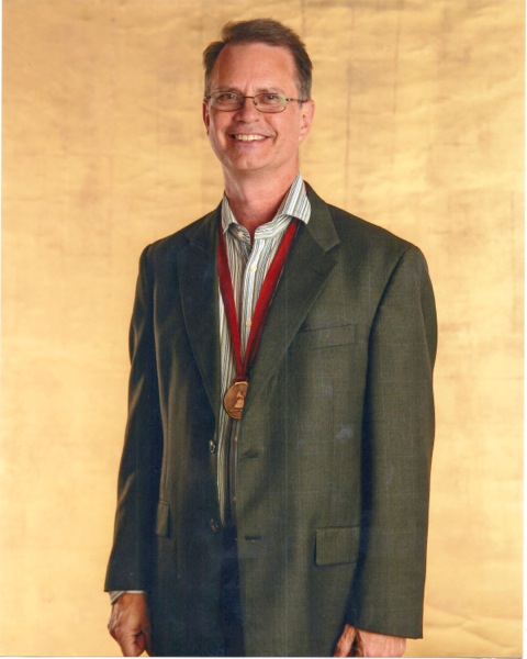 Professor Chuck Owen stands with GRAMMY nomination medal before a gold-colored background