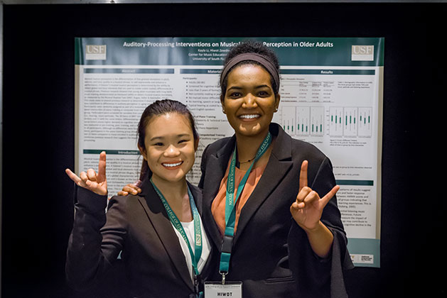Kayla Li and Hiwot Zewdie stand in front of their research poster at the USF Undergraduate Research Symposium giving "horns up" hand gestures