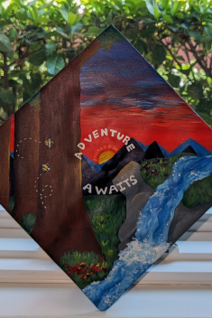 Handpainted grad cap with a forest, mountain, and river scene and the words: "adventure awaits"