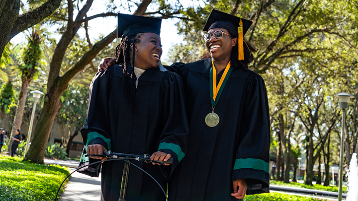 Two USF grads wearing commencement regalia smile and laugh