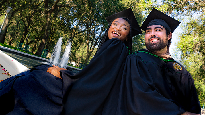 Two USF grads, wearing regalia, smile at each other in front of the fountains on the Tampa campus