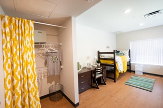 Closet with curtain, a carpet in the middle and bed with yellow comforter on the right