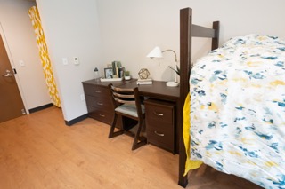 Closet with curtain, brown desk and chair, bed with yellow spread in corner.