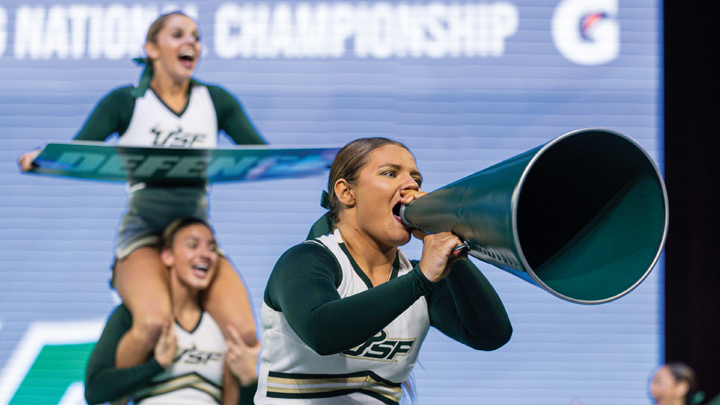 A female USF cheerleadr shows into a large, green megaphone, while a teammate behind her sits on the shoulders of another. They are wearing USF cheer outfits, white bodices with long, dark green sleeves.