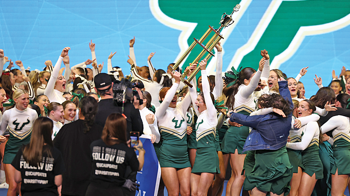 A large crowd of USF cheerleaders in USF uniforms and media jump for joy, holding a giant tropy aloft. They are in a competition space, with a giant USF U-icon displayed on the wall behind them.