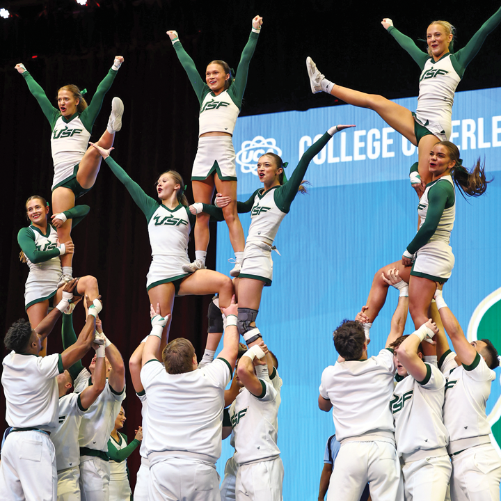 Approximately 16 USF Coed Cheer teammates, wearing USF colors, display a three-person high tower. Male teammates form the 9-person base. Four female cheerleaders form the center, and three female cheerleaders form the top with arms raised high in a V-formation.