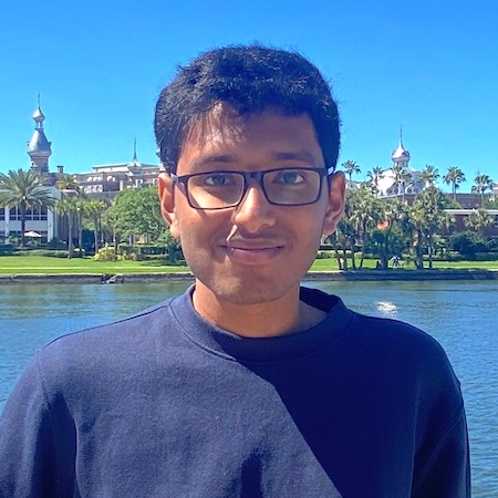 A man with dark hair and glasses smiles in a blue sweater by a lake