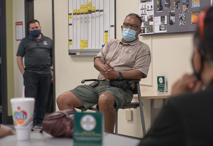Mack sits in a high top chair with a face mask on while discussing his experience to a group of parking employees watching from around the breakroom.