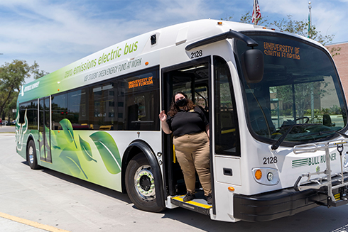 Bull Runner driver waves from the doorway of a parked bus. The bus is lined with large green leafs and says