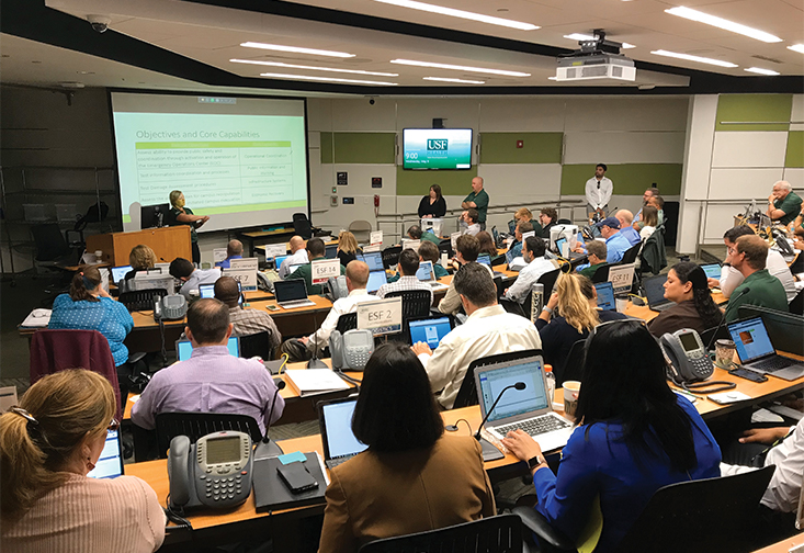 A large lecture hall is filled with staff and computers observing a presentation at the front of the room titled "objectives and core capabilities."