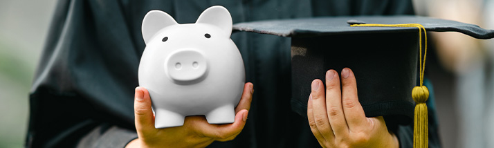 A graduate in a cap and gown holding a piggy bank to symbolize financial savings.