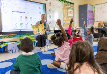 A teacher sits in a classroom holding up a children's book to a group of young participating students sitting on a rug.