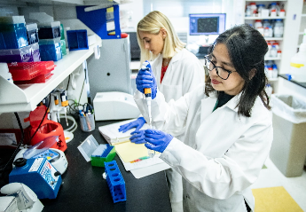 Two students wearing lab coats and blue gloves working together on a project using pipettes and taking down measurements.