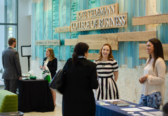 A group of people conversing and networking around tables in the lobby of the Kate Tiedemann College of Business.