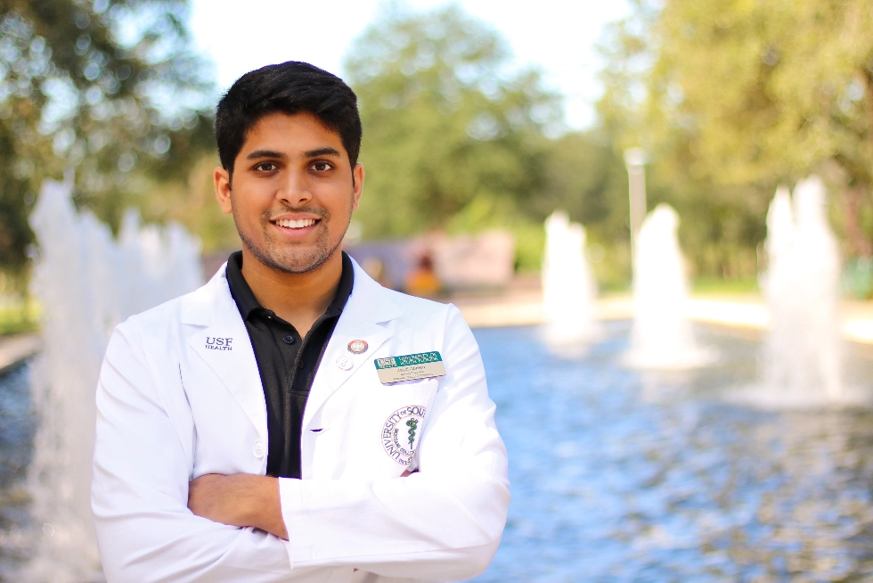A USF student in a white lab coat smiling in front of a fountain on campus.