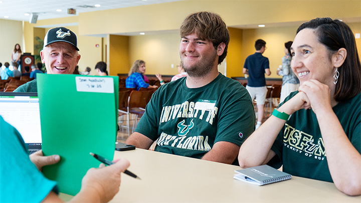 Three individuals are seated at a table in a bright, open space, engaging in conversation and sharing smiles while looking at a person holding a green folder. The setting features a casual atmosphere with other people in the background, indicating a welcoming community event.