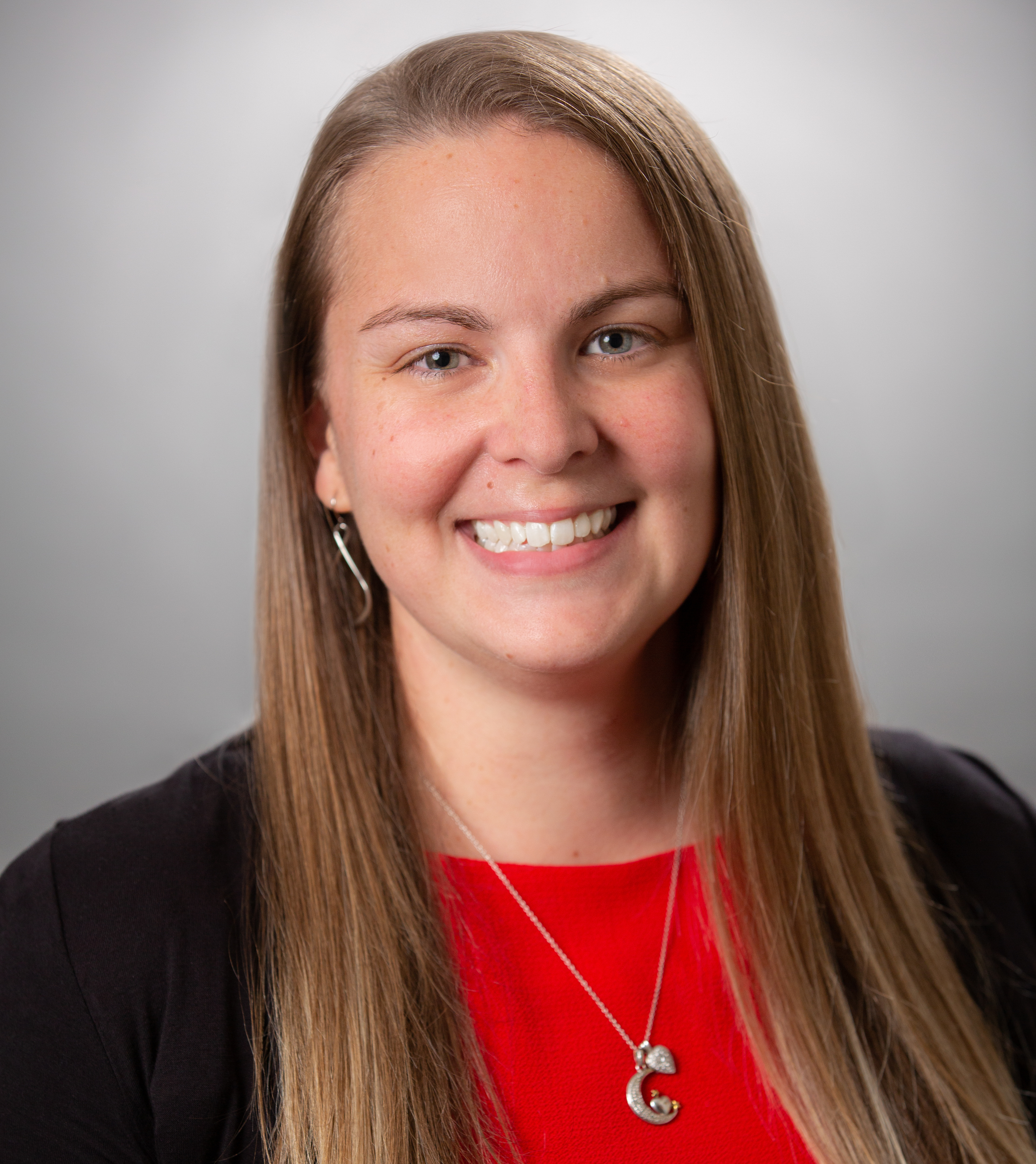 Professional headshot of a smiling woman with long light brown hair, wearing a red top, black blazer, silver necklace, and earrings against a gray background.