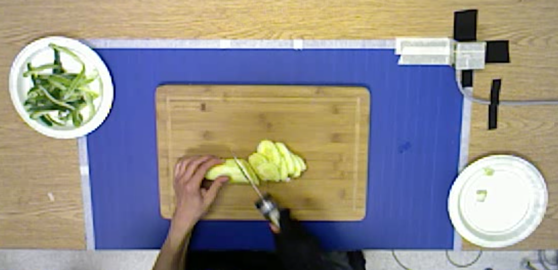 A person slices yellow squash on a wooden cutting board, using a knife, while green onions are placed in a bowl to the left, and a white plate sits to the right on a purple surface. The setting appears to be a kitchen workspace with a wooden background.