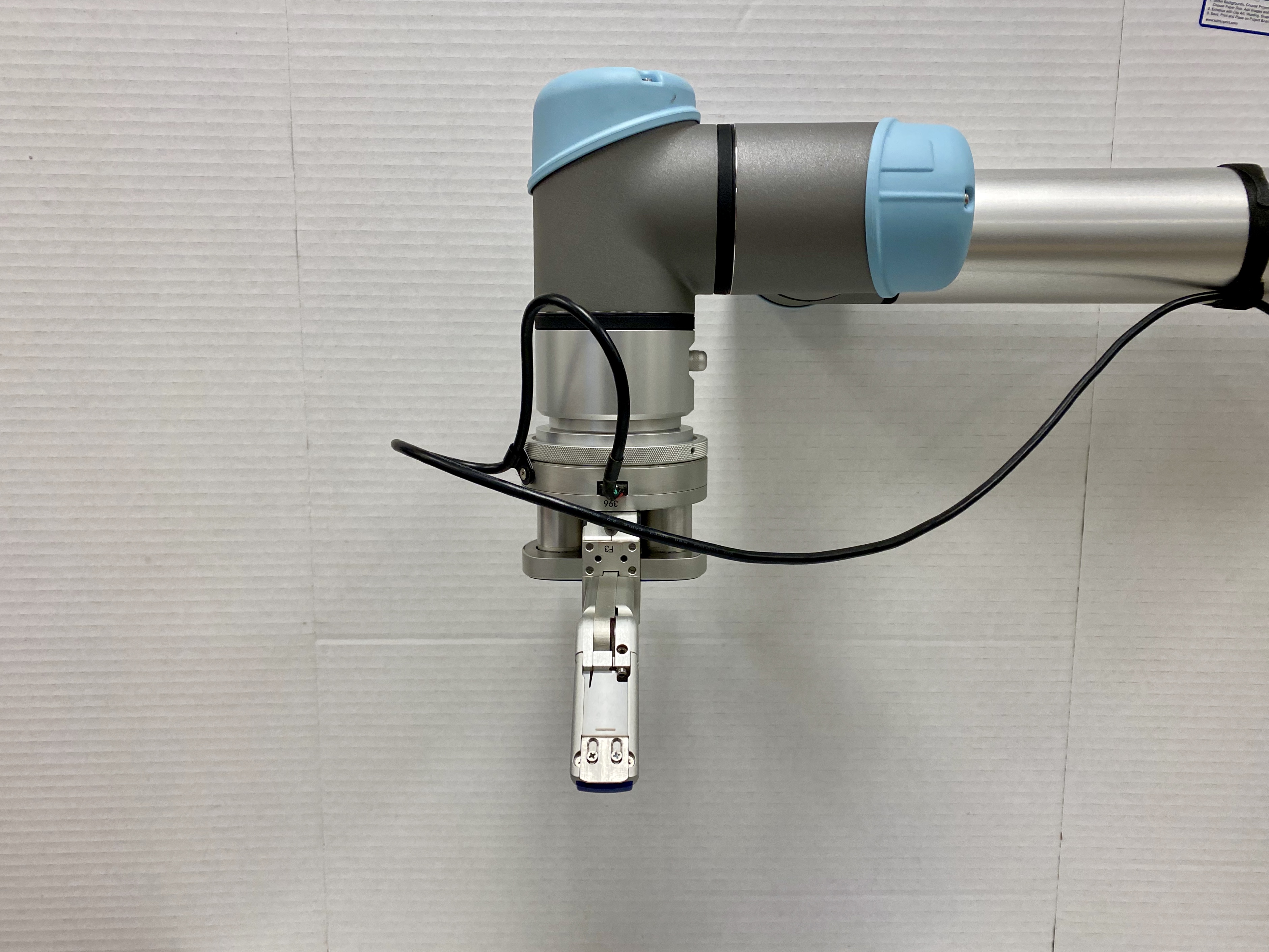 A robotic arm with a light blue and silver design is shown against a plain white background, featuring a clamp-like gripper at the end. Wires are connected to the arm, indicating it's likely part of a technological setup or industrial automation system.
