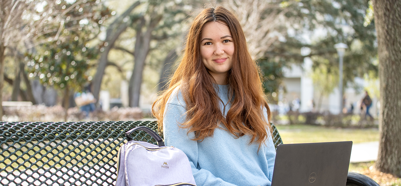 Anne Utegen sits on a bench under a shaded area holding her laptop.