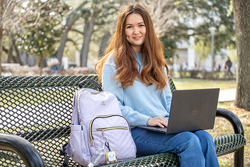 Anne Utegen sits on a bench under a shaded area holding her laptop.