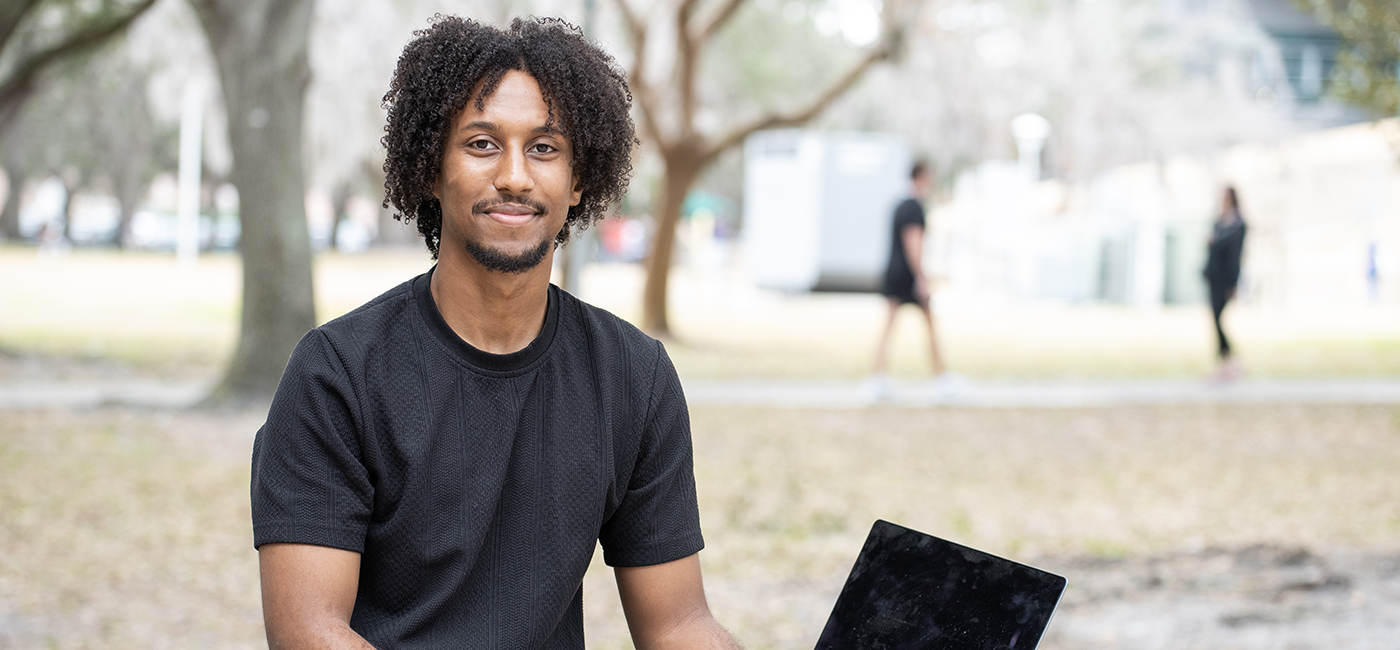 A portrait of Tesga Teshome sitting next to a computer under shade from trees