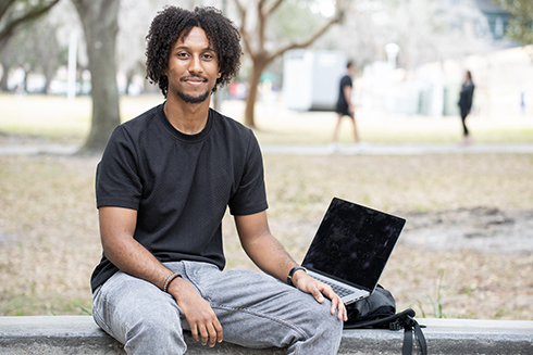 A portrait of Tesga Teshome sitting next to a computer under shade from trees