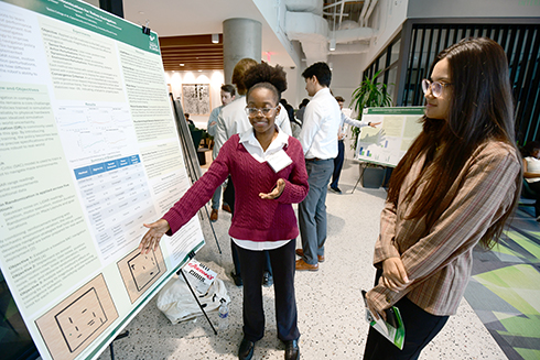 A young woman in a burgundy sweater enthusiastically presents her research at a poster session, gesturing towards her display, while another woman in glasses listens attentively; the setting features modern architecture and a group of attendees mingling in the background.