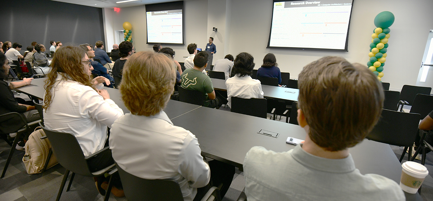 An audience of diverse individuals sits in a modern conference room, attentively watching a presenter in front of a large screen displaying a research overview, with colorful decorations in the background. The setting features tables and chairs, creating an engaging environment for discussion and learning.