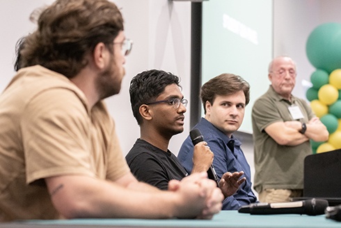 A panel discussion features three speakers, one actively speaking into a microphone, while the others listen intently, with a backdrop of colorful balloons and a presentation screen in a modern setting.