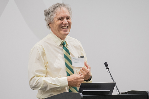 A man with curly hair and a broad smile stands behind a podium, wearing a light yellow shirt with a green and gold tie, as he gestures animatedly while speaking to an audience in a modern conference setting.