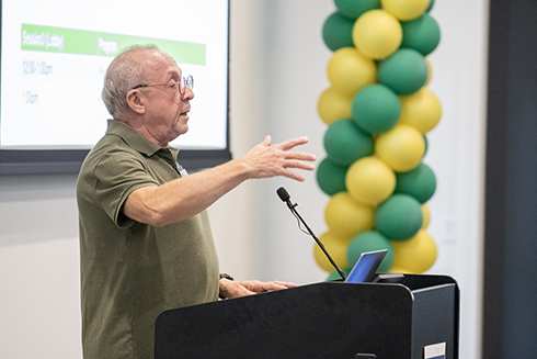 An older man with glasses gestures while speaking at a podium in front of a screen displaying event details, alongside a colorful balloon column in green and yellow.