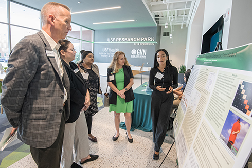 A group of four individuals, including a woman in a black dress, stands attentively around a presentation board in a modern, brightly lit room, while a presenter gestures to the board, engaging her audience at the USF Research Park.