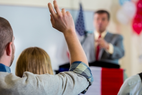 Person with raised hand in audience facing speaker at a podium (Photo source: Canva)