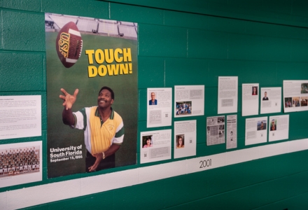 A portion of the timeline covering the late 1990s and early 2000s includes a poster (purchased on eBay) of Lee Roy Selmon commemorating the approval of the football team in 1995; the exhibit notes that Professor Emeritus of Anthropology Dr. Curtis Wiencker was a member of the committee that brought football to campus. (Photo by Corey Lepak)