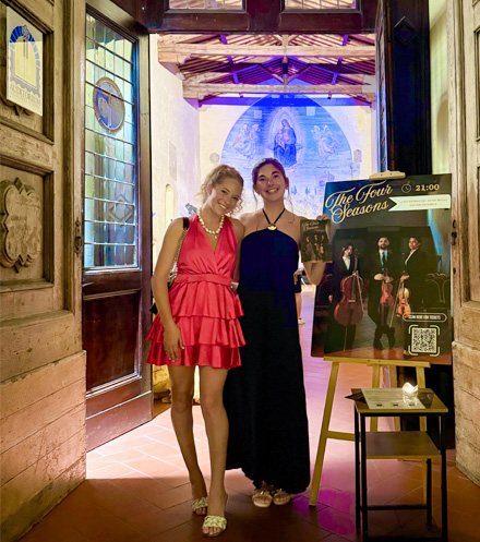 Two people standing in the doorway of an ornate historic building, one in a bright pink dress and one in a dark dress, beside an easel displaying a poster for a musical event.
