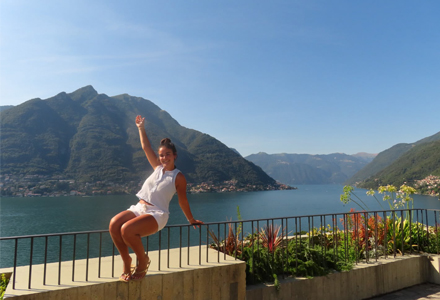 Person sitting on a stone ledge overlooking a large lake surrounded by tall green mountains, raising one arm and smiling on a sunny day with flowers in the foreground.