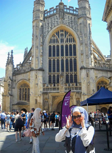 Visitors stand in front of a large Gothic-style cathedral on a sunny day, with people walking nearby and signage and a canopy tent visible near the entrance.