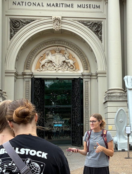 Group of people gathered outside the entrance of the National Maritime Museum, standing near an ornate archway with carved details and signage, while someone in the foreground speaks to them.