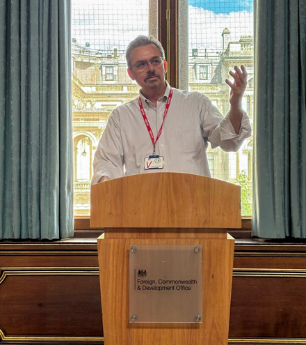 A male professor in a white collared shirt and red lanyard presenting at a podium at the Foreign Commonwealth & Development Office in London.