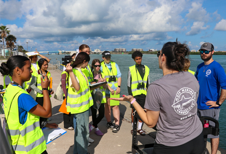 A group of high school students wearing yellow safety vests stand near the water with clipboards and cameras while a marine science instructor speaks to them. Palm trees, a bridge, and buildings line the background under a partly cloudy sky