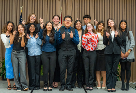 Thirteen people stand together on a stage in front of a curtain backdrop with two American flags. They pose for a group photo, smiling and making celebratory hand gestures, dressed in semiformal attire.