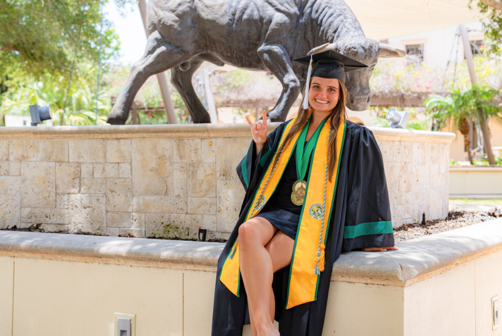 Britlee Yant in USF cap and gown holding up the Go Bulls hand sign with a bull statue in the background