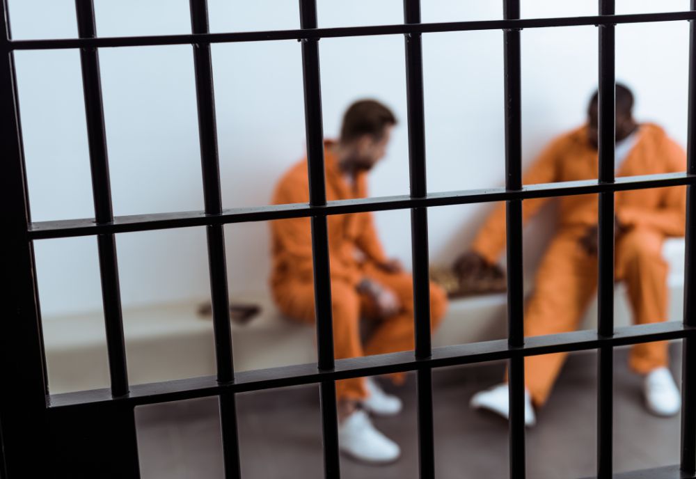 two inmates in orange jumpsuits sit behind jail bars