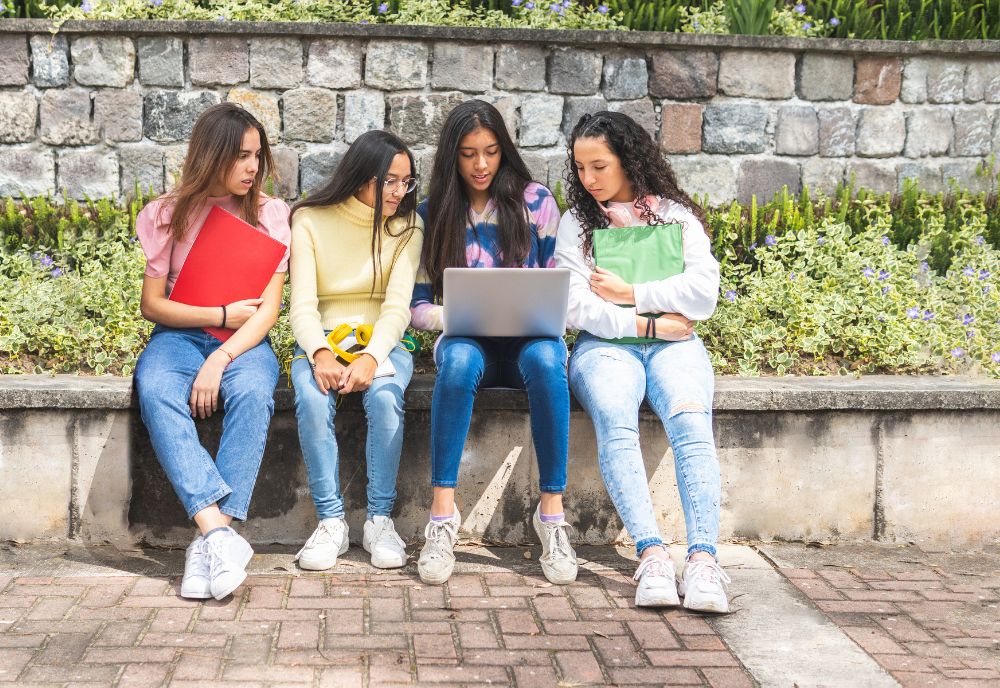 four young women seated on a low wall bench look at laptop held by one of the women (photo by Adobe Stock)
