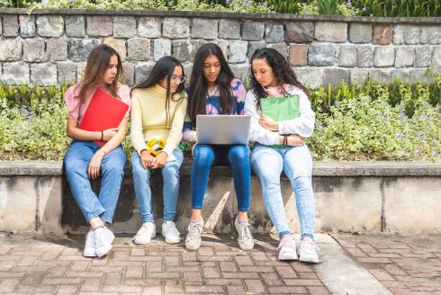four young women seated on a low wall bench look at laptop held by one of the women (photo by Adobe Stock)