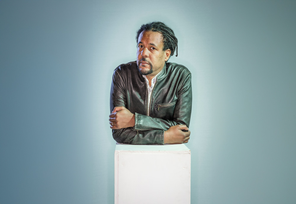 Colson Whitehead wearing a black leather jacket with arms crossed on a stand.
