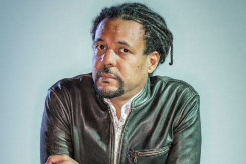 Colson Whitehead wearing a black leather jacket with arms crossed on a stand.