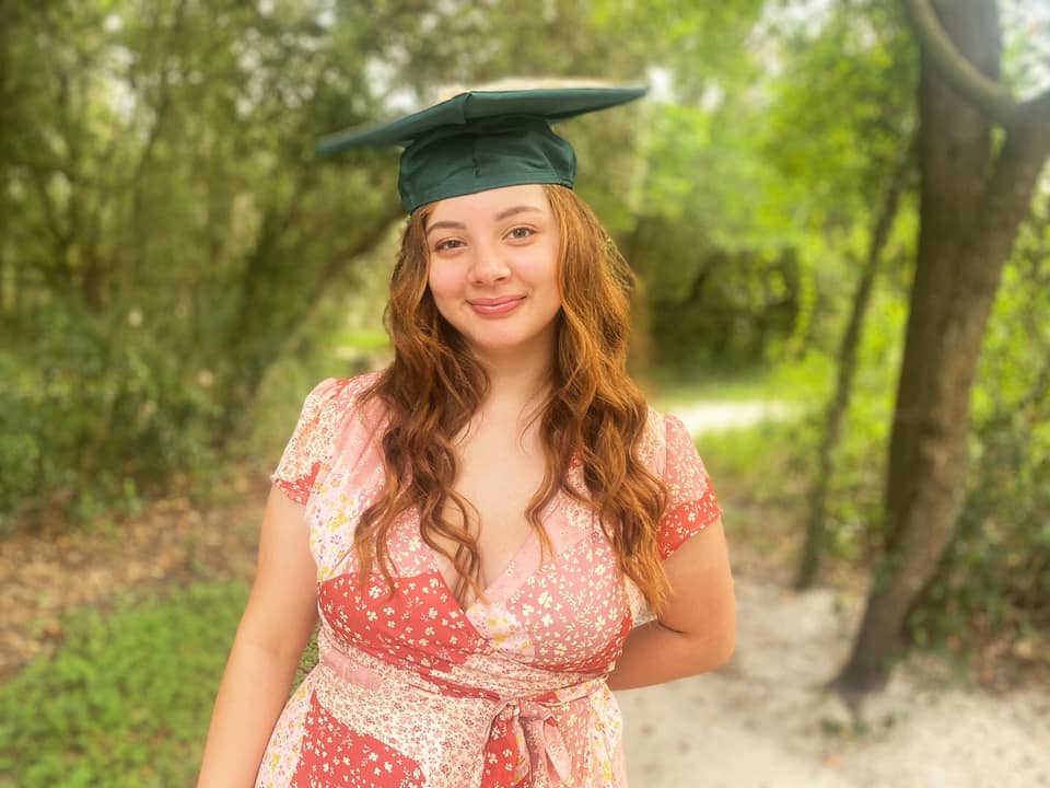 A white woman with red hair wearing a pink and white summer dress and a USF green graduation cap